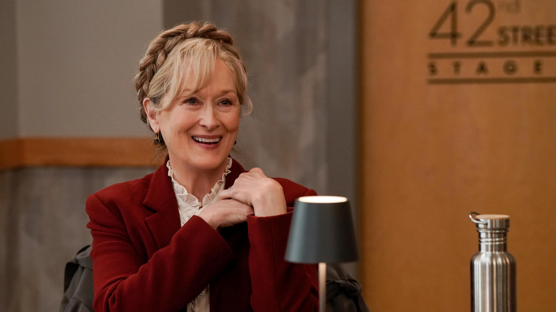 A woman with gray-blond hair wrapped in a Dutch braid, wearing a red blazer and looking delighted; still of Meryl Streep from "Only Murders in the Building"