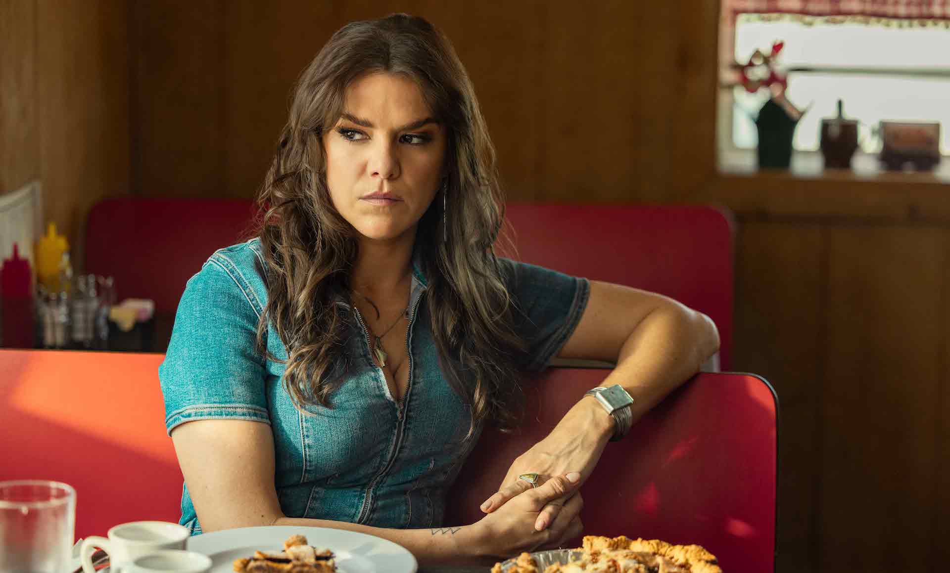 A woman with long, wavy brown hair wearing a denim zip-up top and sitting in a red diner booth in front of a whole pie; still from "Reservation Dogs"